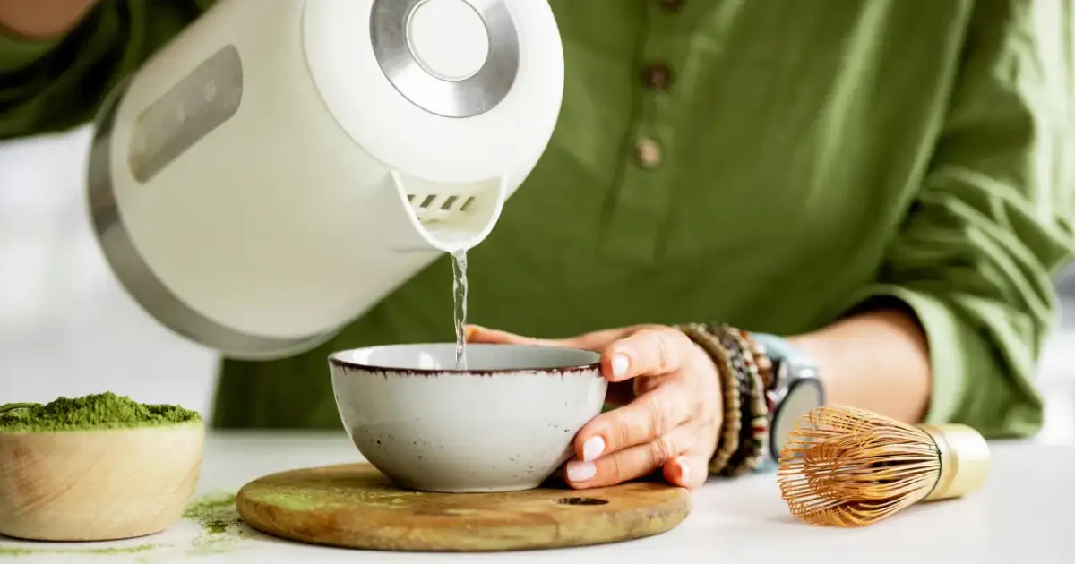 Przygotowanie napoju. Gorąca woda z czajnika leje się do miseczki z zielonym proszkiem. To właśnie co to jest matcha, przygotowywana z pomocą bambusowej miotełki.