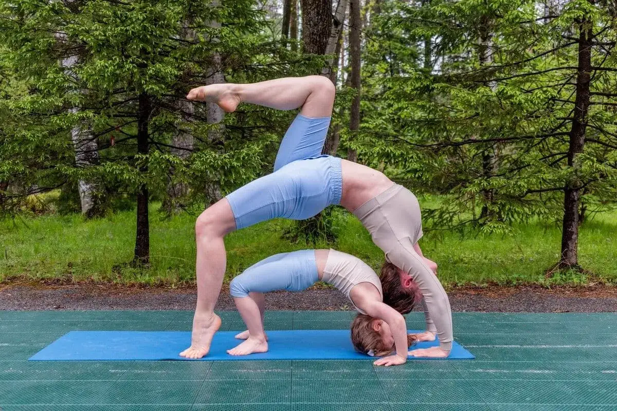 Dwie osoby wykonują ćwiczenia jogi w pozycji mostka na macie na tle lasu.