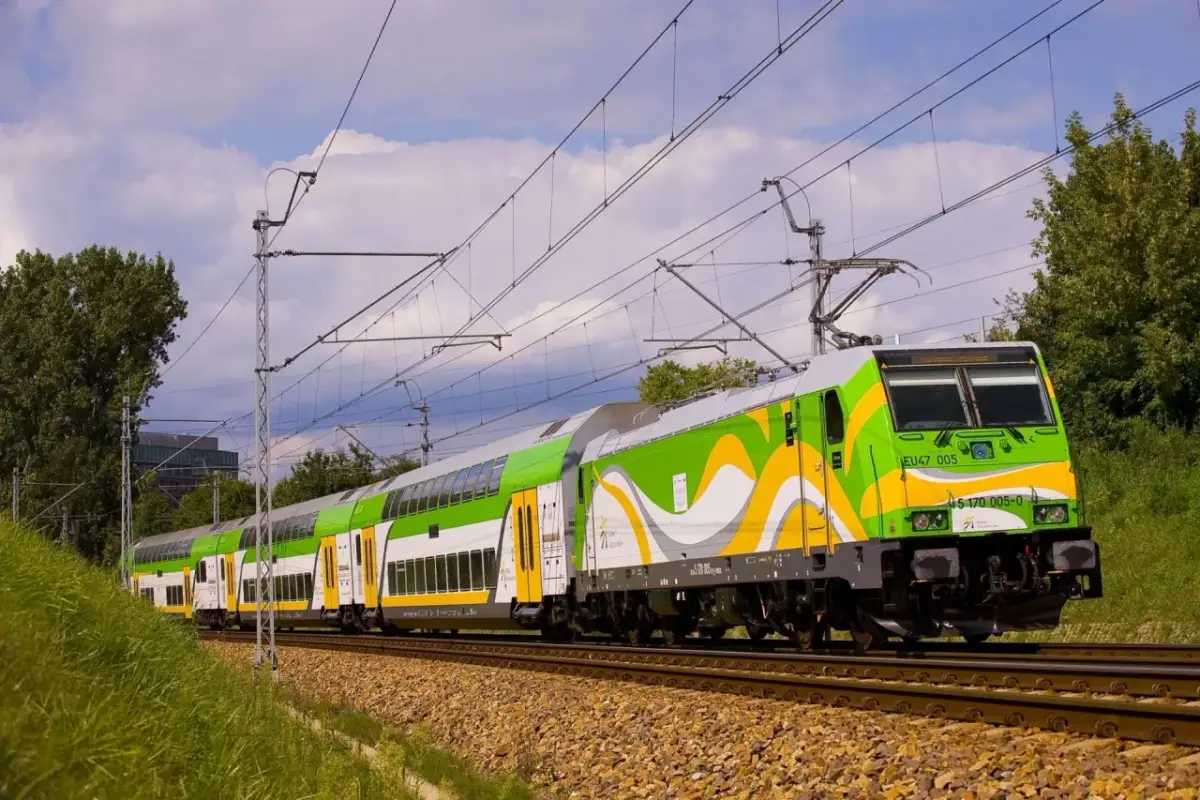 Zielono-biały pociąg z żółtymi akcentami na torach, pod niebem z chmurami.