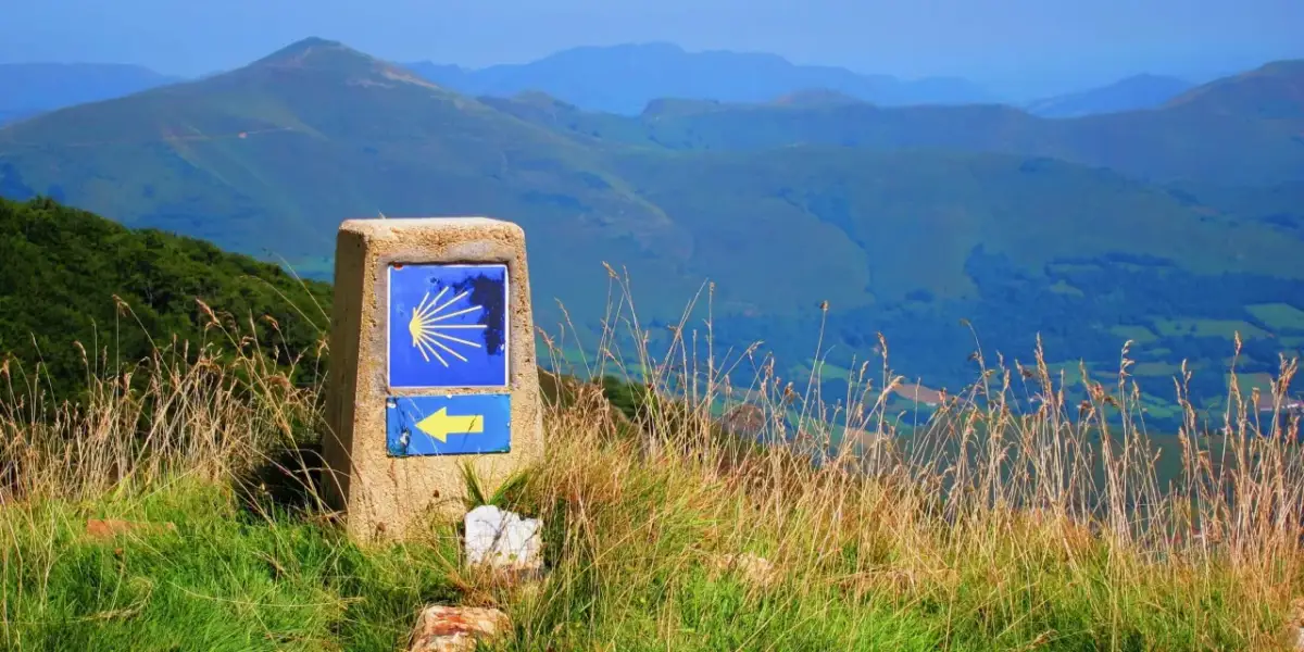 Kamień z symbolem muszli i żółtą strzałką, wskazujący drogę na Santiago de Compostela szlak z Polski. W tle góry.