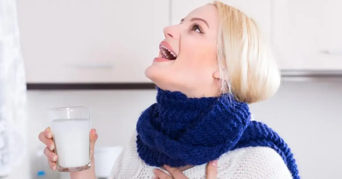 Kobieta w niebieskim szaliku wykonuje płukanie gardła solą, trzymając szklankę z płynem.