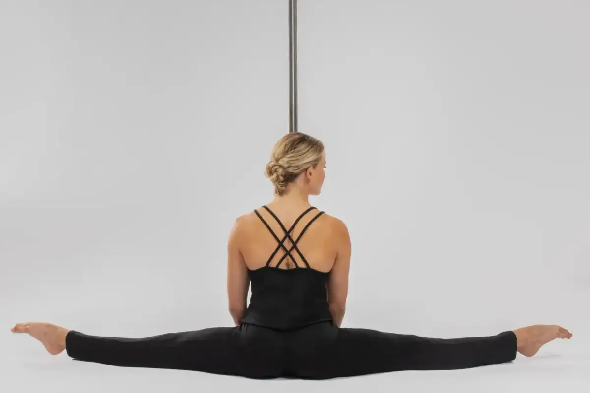 Kobieta w czarnym stroju siedzi w szpagacie przy rurze do pole dance.