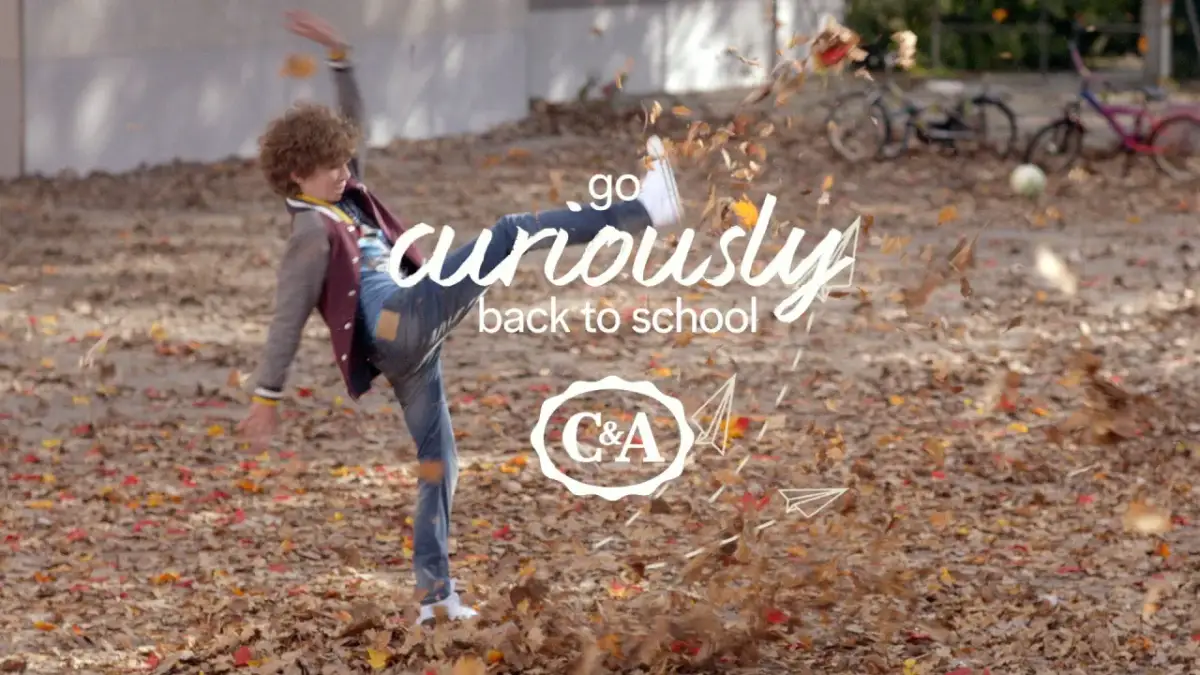 Chłopiec w jesiennych ubraniach kopie liście, z napisem "go curiously back to school" i logo C&A.