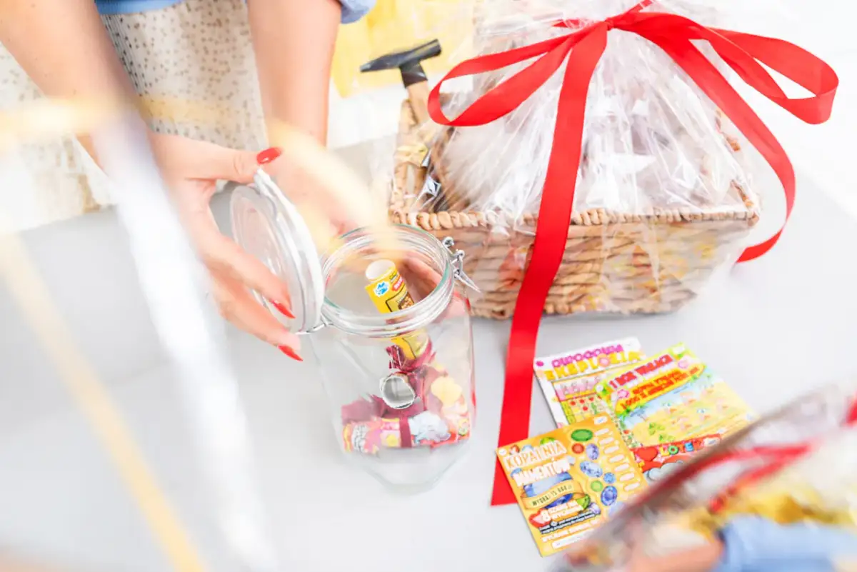 Ręce z czerwonymi paznokciami wkładają zdrapki do słoika, przygotowując je do zapakowania na prezent. Obok kosz z upominkami.