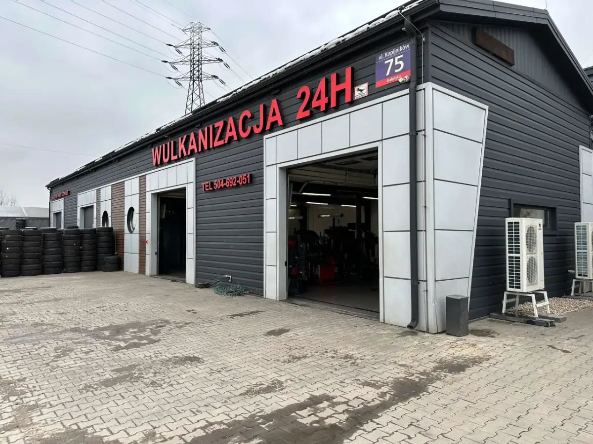 Budynek wulkanizacji z napisem "Wulkanizacja 24H", stosy opon i otwarte bramy garażowe.