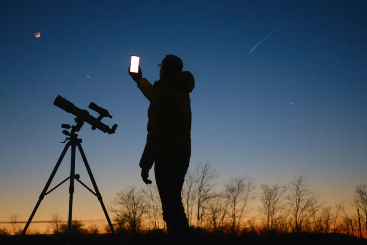 Sylwetka osoby z telefonem i teleskopem na tle nieba z zaćmieniem Księżyca i smugą po samolocie.