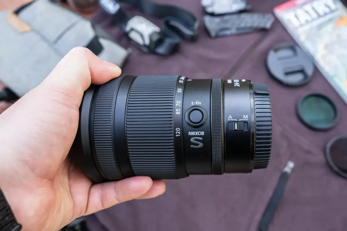 Dłoń trzyma obiektyw Nikkor S 24-120mm, idealny do pełnej klatki Nikon. W tle akcesoria fotograficzne.