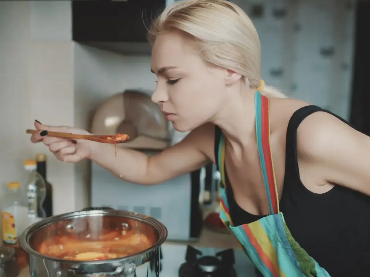 Kobieta w kolorowym fartuchu wącha z łyżki gotującą się zupę. Może to być część przepisu jak zahartować jogurt naturalny.