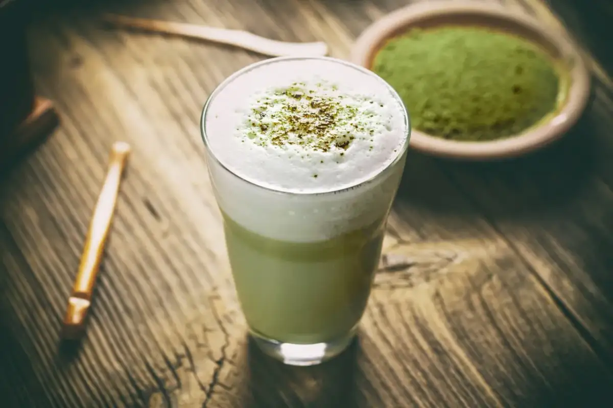 Szklanka z zielonym matcha latte z pianką, obok miseczka z proszkiem matcha i bambusowa trzepaczka.