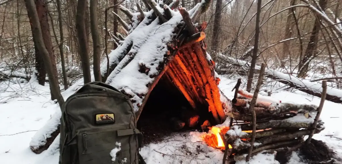 Jak zbudować szałas zimą? W lesie, wśród śniegu, płonie ognisko ogrzewające prowizoryczne schronienie z gałęzi. Obok stoi plecak.