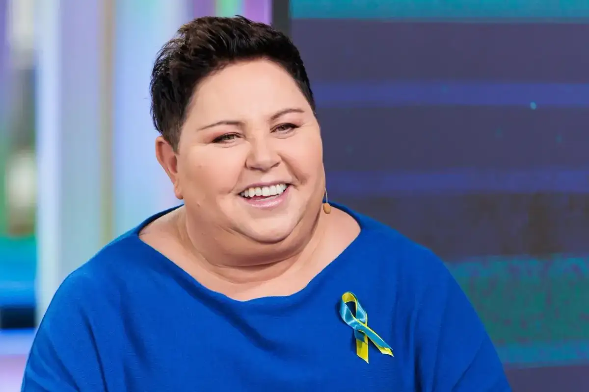 Uśmiechnięta Dorota Wellman w niebieskim swetrze z przypinką w barwach Ukrainy. Jej wykształcenie i doświadczenie pomagają jej w pracy.