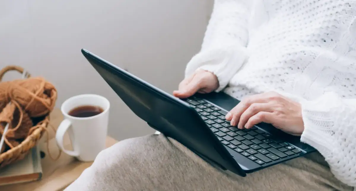 Osoba w białym swetrze pisze na laptopie, obok kubek z kawą i koszyk z włóczką.