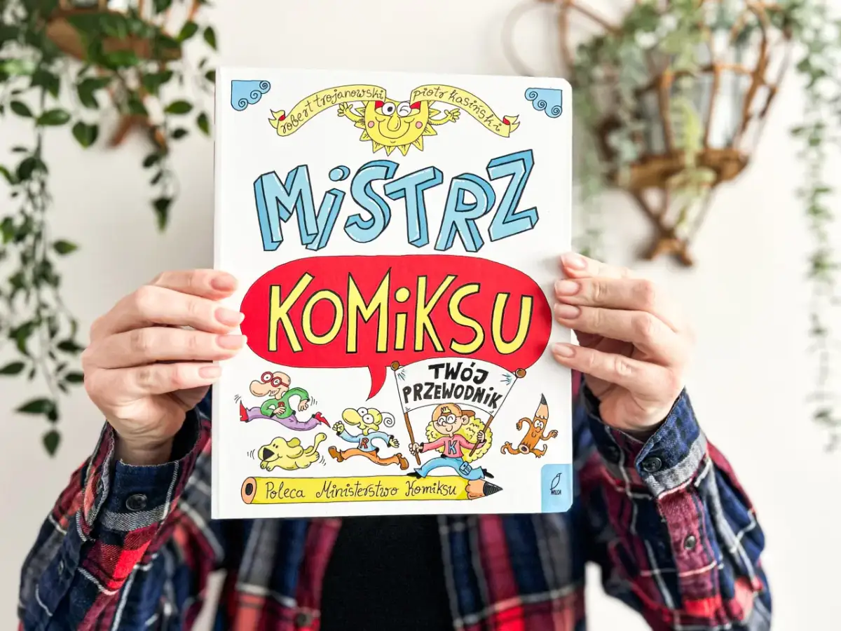 Okładka książki "Mistrz Komiksu" z rysunkami postaci i słońca, trzymana przez ręce.