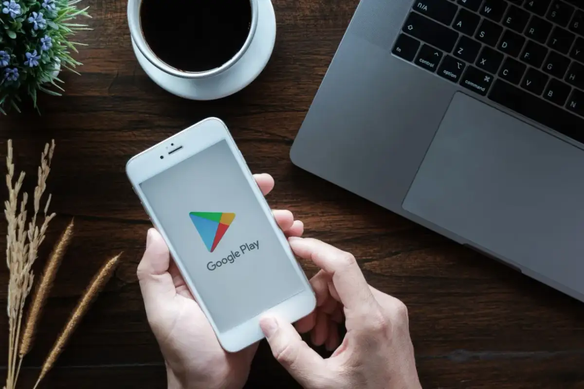 Ręce trzymają smartfon z logo Google Play, gotowe by dowiedzieć się, jak zaktualizować usługi Google Play. Obok laptop i kawa.