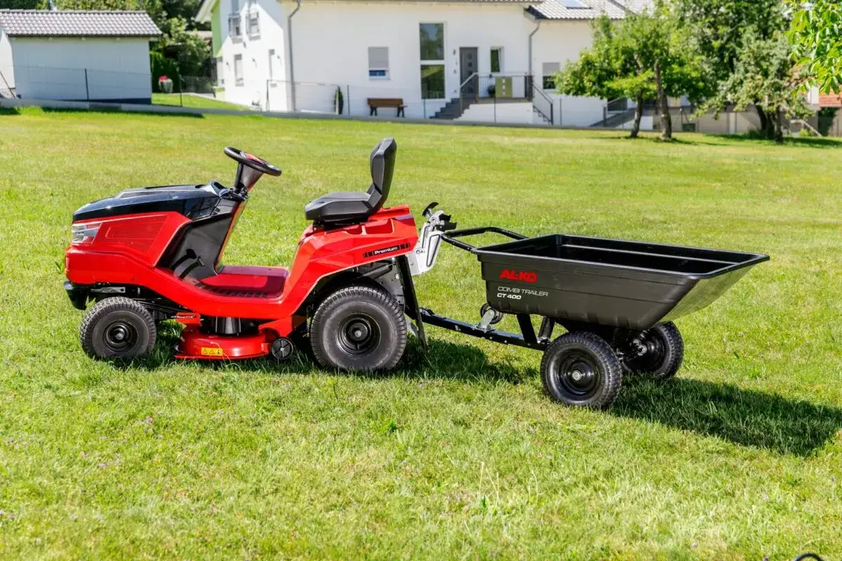 Czerwony traktorek ogrodowy z czarną przyczepką do traktorka na zielonej trawie, gotowy do pracy.