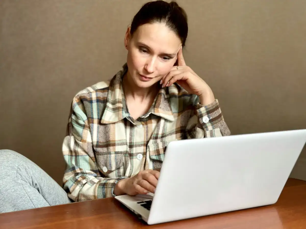 Kobieta w kratę siedzi przy laptopie, zamyślona, z ręką na skroni.