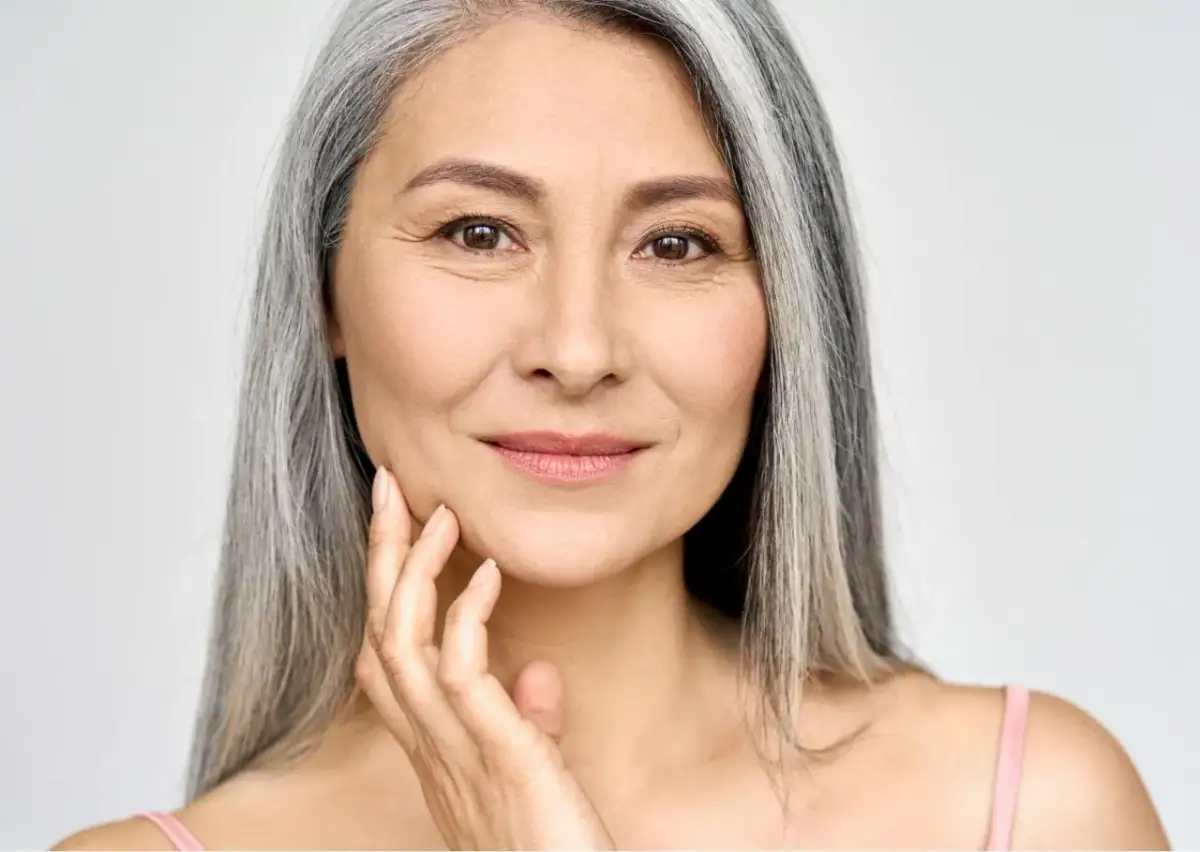 Portret uśmiechniętej kobiety z siwymi włosami, dotykającej dłonią policzka.