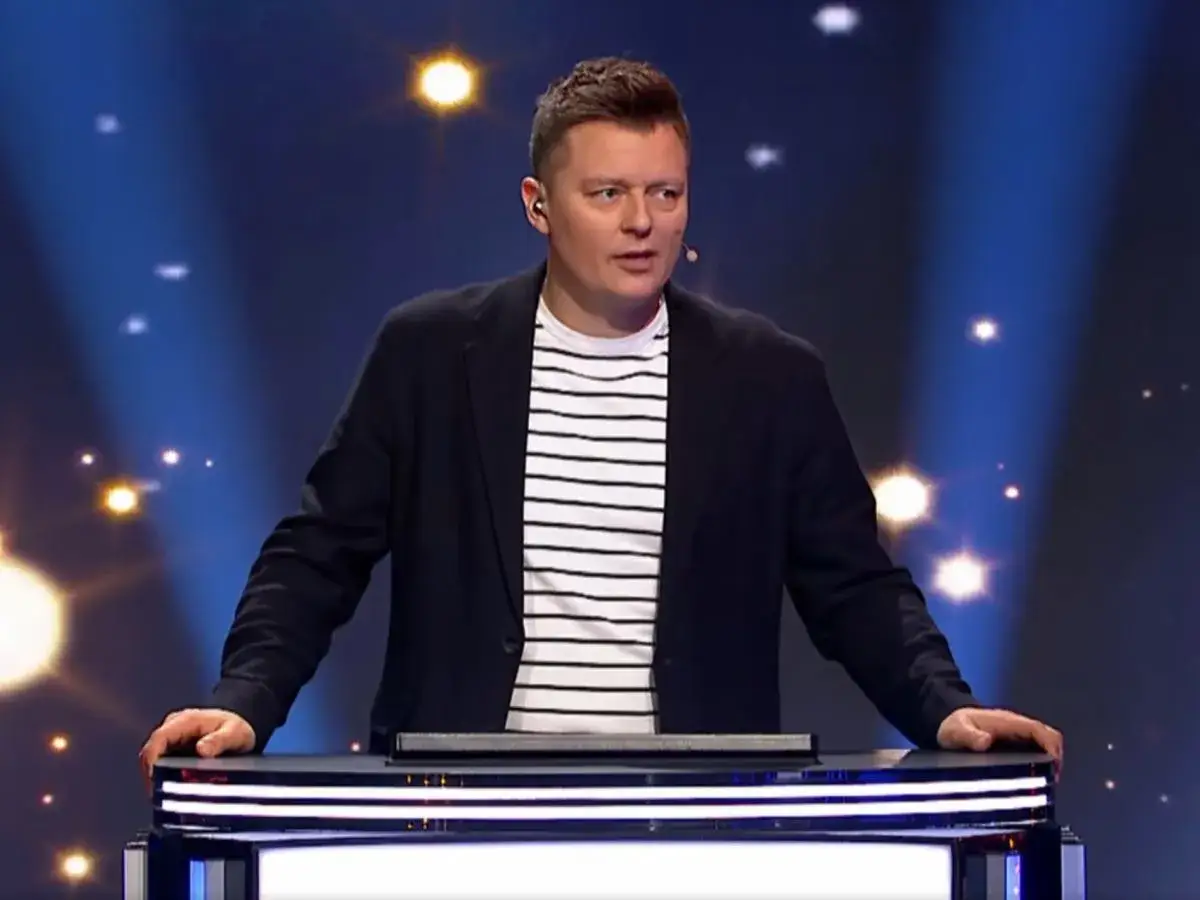 Prowadzący program "Jaka to melodia?" w eleganckim stroju, zastanawia się, ile zarabia zawodowy gracz w Jaka to melodia za jeden odcinek.