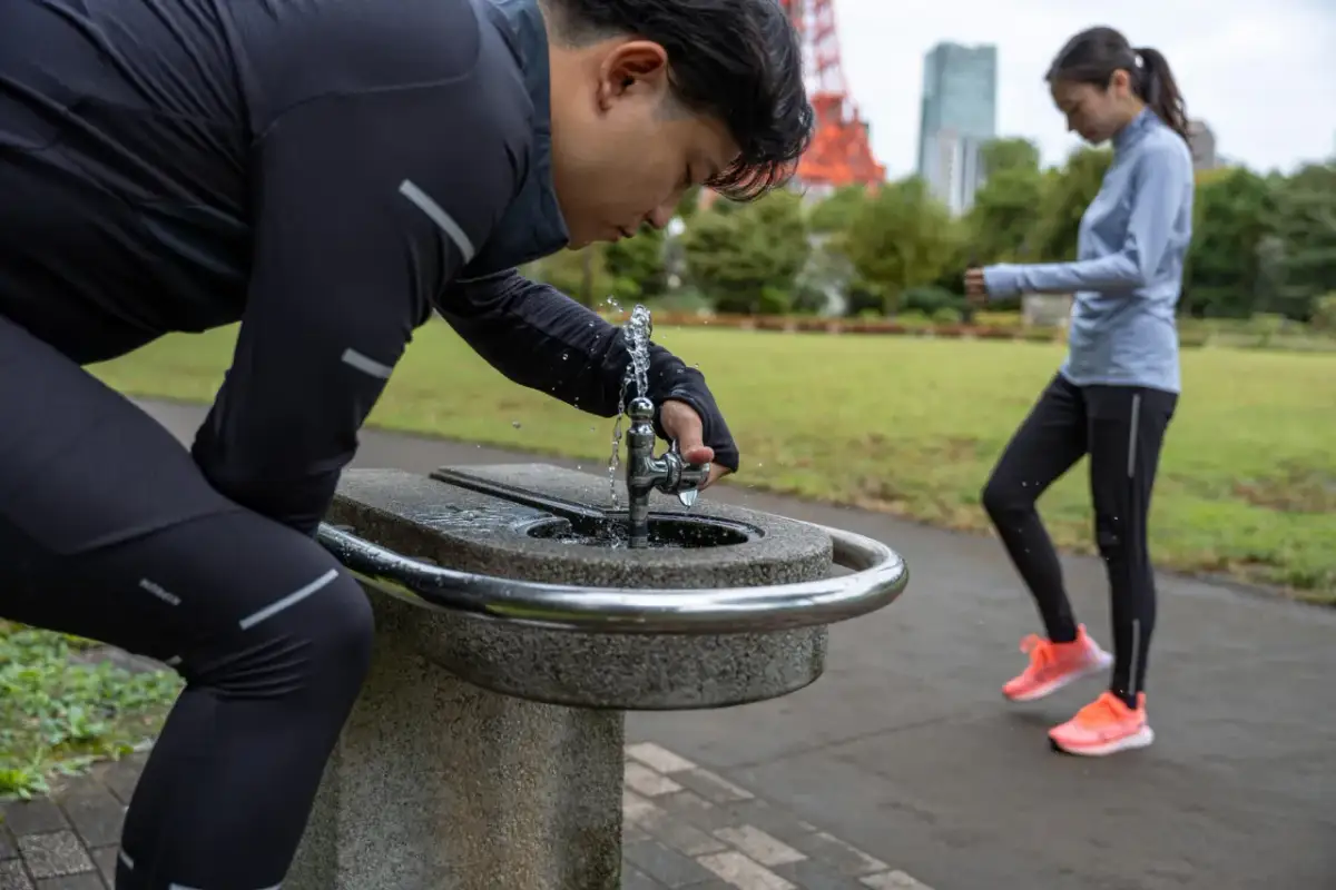 Mężczyzna po bieganiu pije wodę, by złagodzić objawy astmy wysiłkowej. W tle kobieta kontynuuje trening.