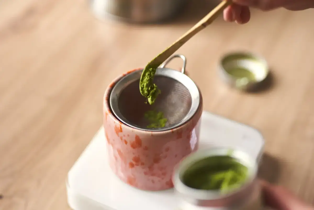 Przygotowanie idealnej matchy: przesiewanie zielonego proszku bambusową łyżeczką do różowego czajniczka. To klucz do pytania, jaka matcha najlepsza.