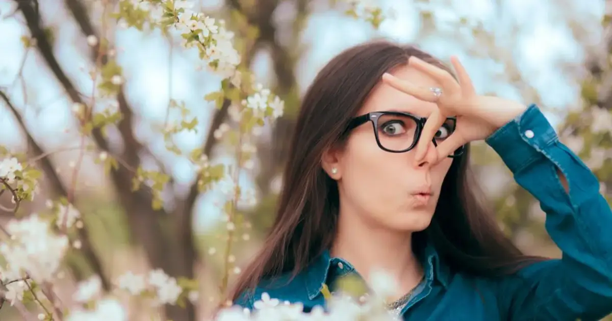 Kobieta w okularach zaciska nos, otoczona kwitnącymi drzewami. Wiosna, piękna, ale dla niej to udręka z powodu kataru siennego.