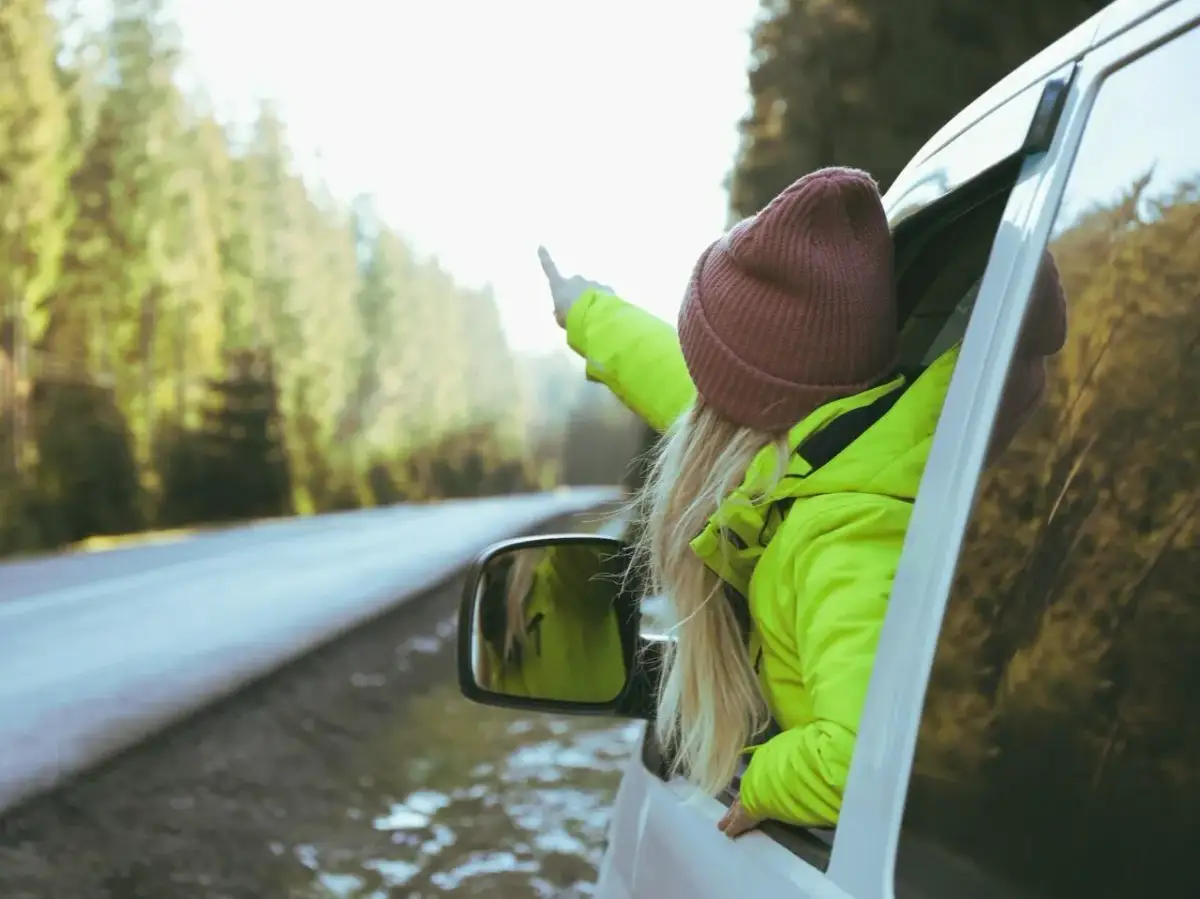 Dziewczyna w jaskrawozielonej kurtce i bordowej czapce wystawia rękę przez okno samochodu, wskazując palcem. Idealne dodatki do zielonej kurtki na podróż!