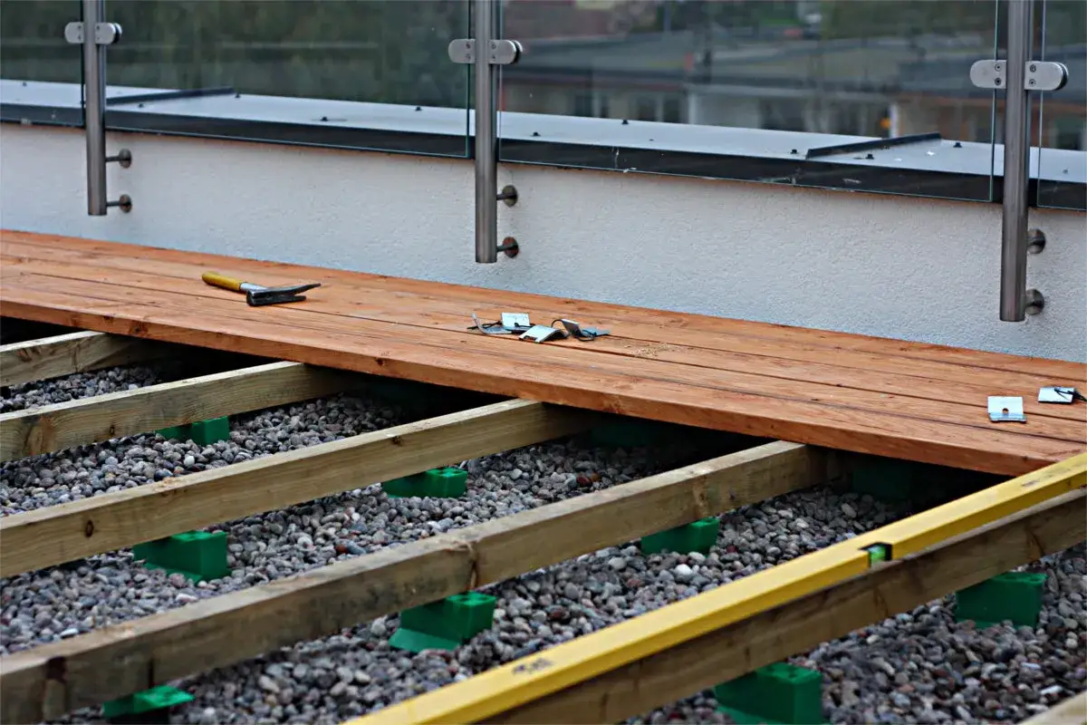 Budowa tarasu drewnianego: deski, legary na zielonych podporach, żwir i młotek. Zastanawiasz się, ile kosztuje taras drewniany?