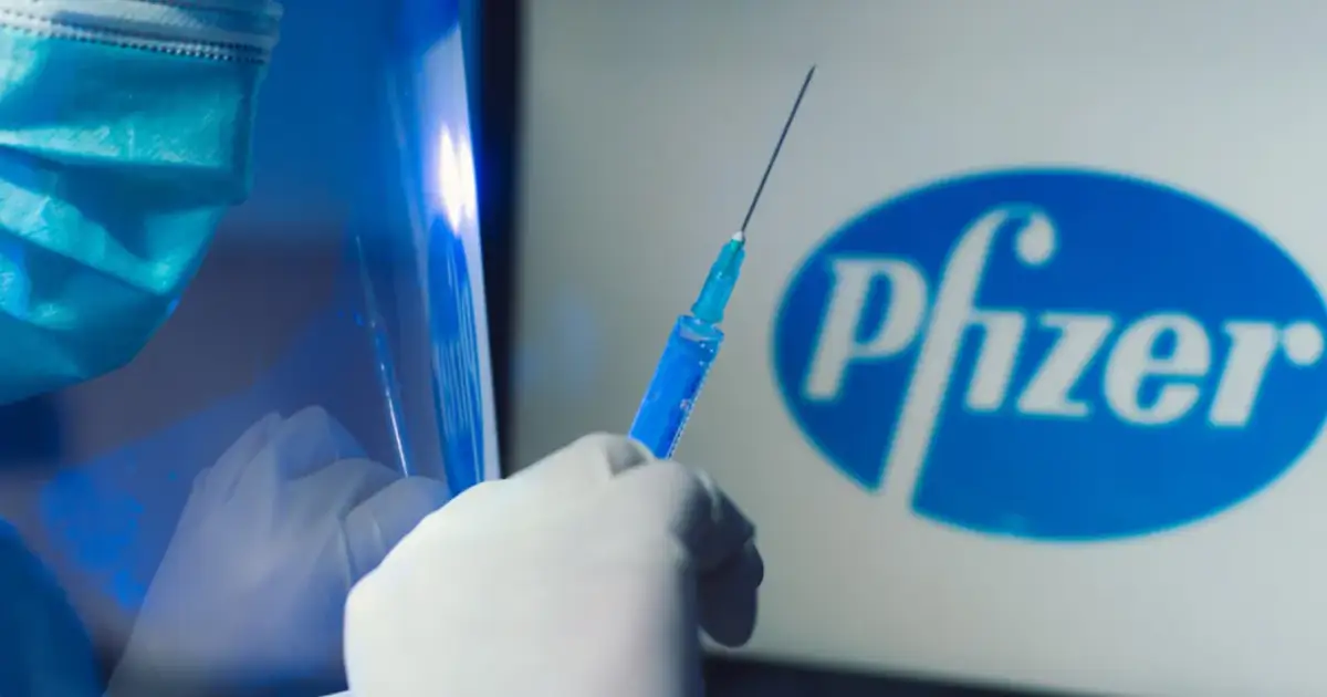 Ręka w rękawiczce trzyma strzykawkę z niebieskim płynem na tle logo Pfizer. Dyskusja o skutkach ubocznych szczepionki Pfizer trwa.