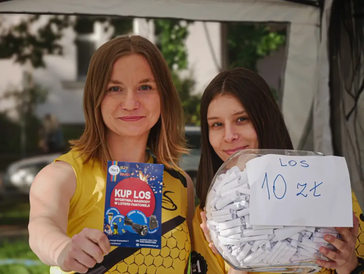 Dwie kobiety w żółtych koszulkach trzymają ulotkę "Kup los" i słoik z losami na loterię fantową.