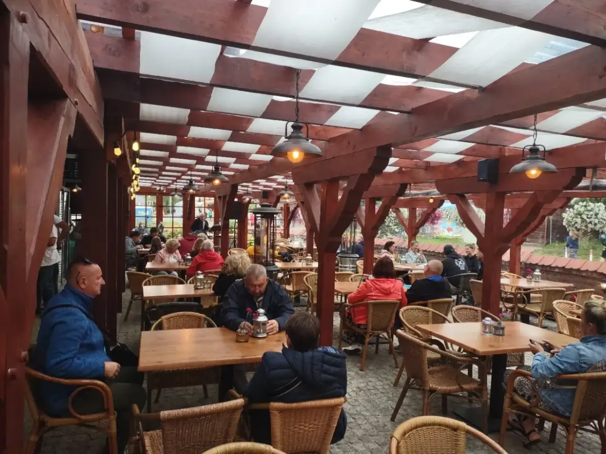Zadaszony ogródek restauracyjny z drewnianymi belkami, lampami i siedzącymi przy stołach ludźmi.