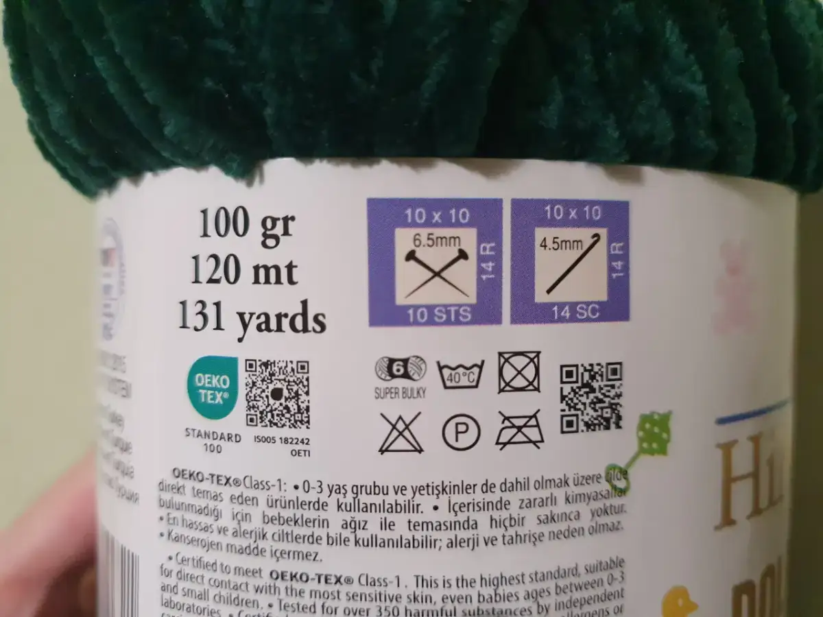 Kłębek ciemnozielonej, puszystej włóczki z etykietą informującą o wadze, metrażu i zaleceniach.