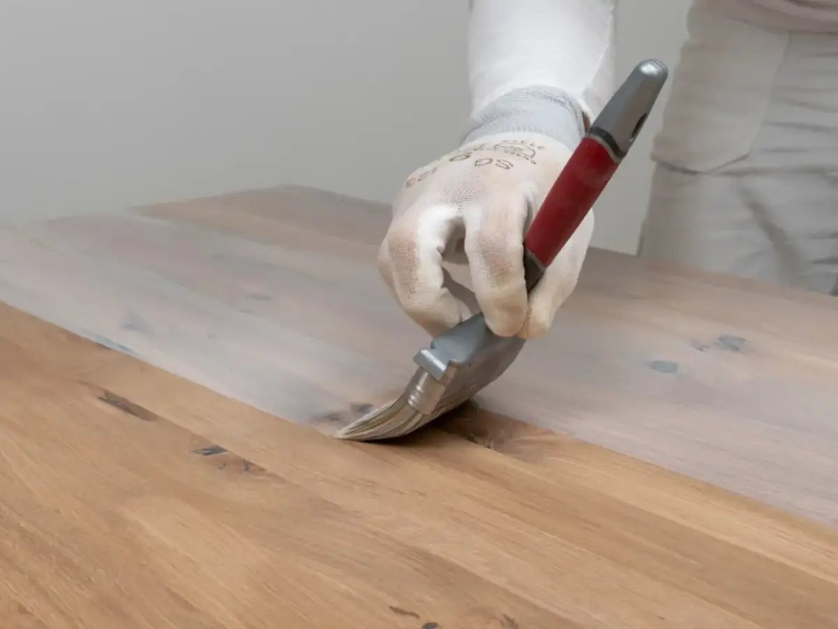 Ręka w rękawiczce nakłada biały preparat na drewniany blat, pokazując, jak usunąć wosk z drewna.