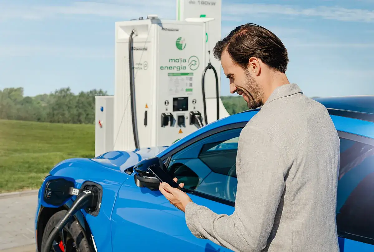 Mężczyzna z telefonem ładuje niebieski samochód elektryczny przy stacji ładowania.