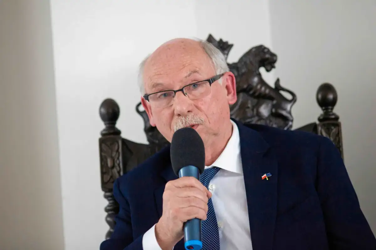 Starszy mężczyzna w okularach i garniturze przemawia do mikrofonu. Na klapie marynarki ma przypinkę z flagami Polski i UE, symbolizującą jego poparcie dla tych, kto głosował za zielonym ładem.