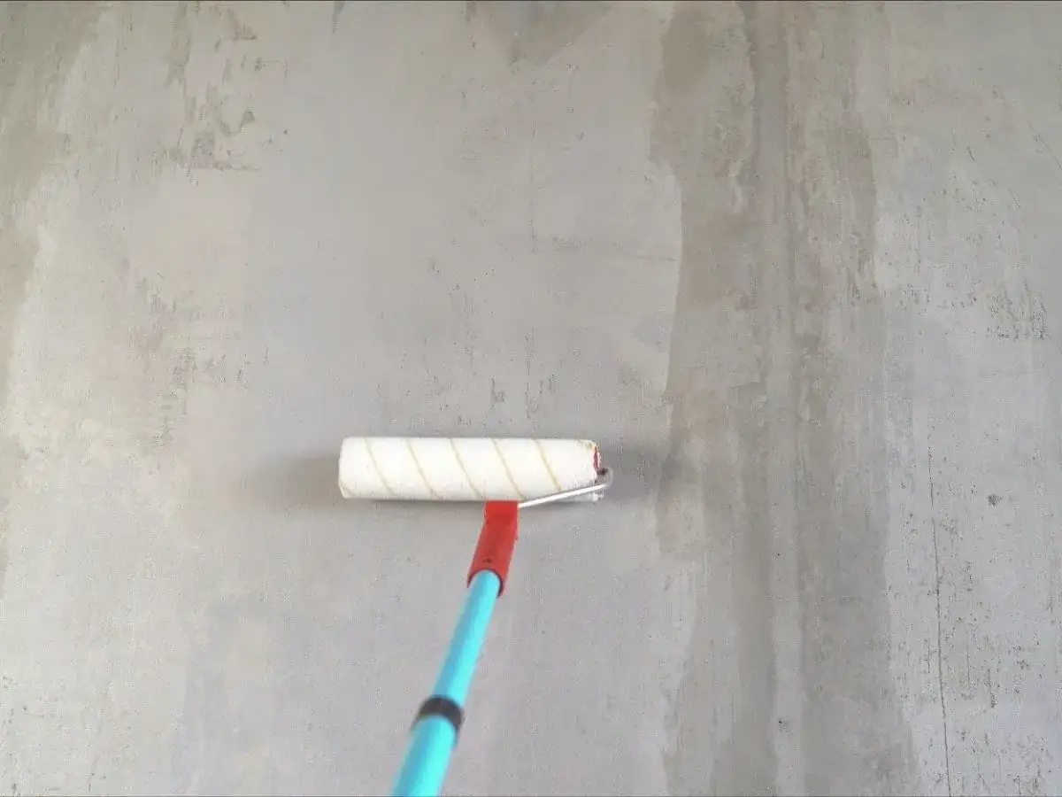 Gruntowanie ścian i sufitów przed tynkowaniem. Wałek malarski z niebieskim trzonkiem nanosi preparat na szarą, betonową powierzchnię.