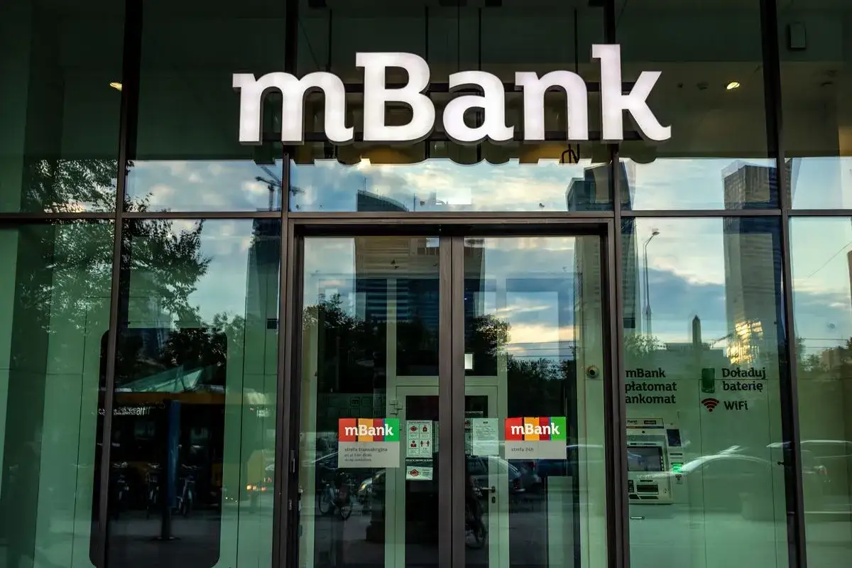 Logo mBank nad wejściem do placówki. Na drzwiach naklejki informujące o usługach, np. jak zablokować kartę mbank.