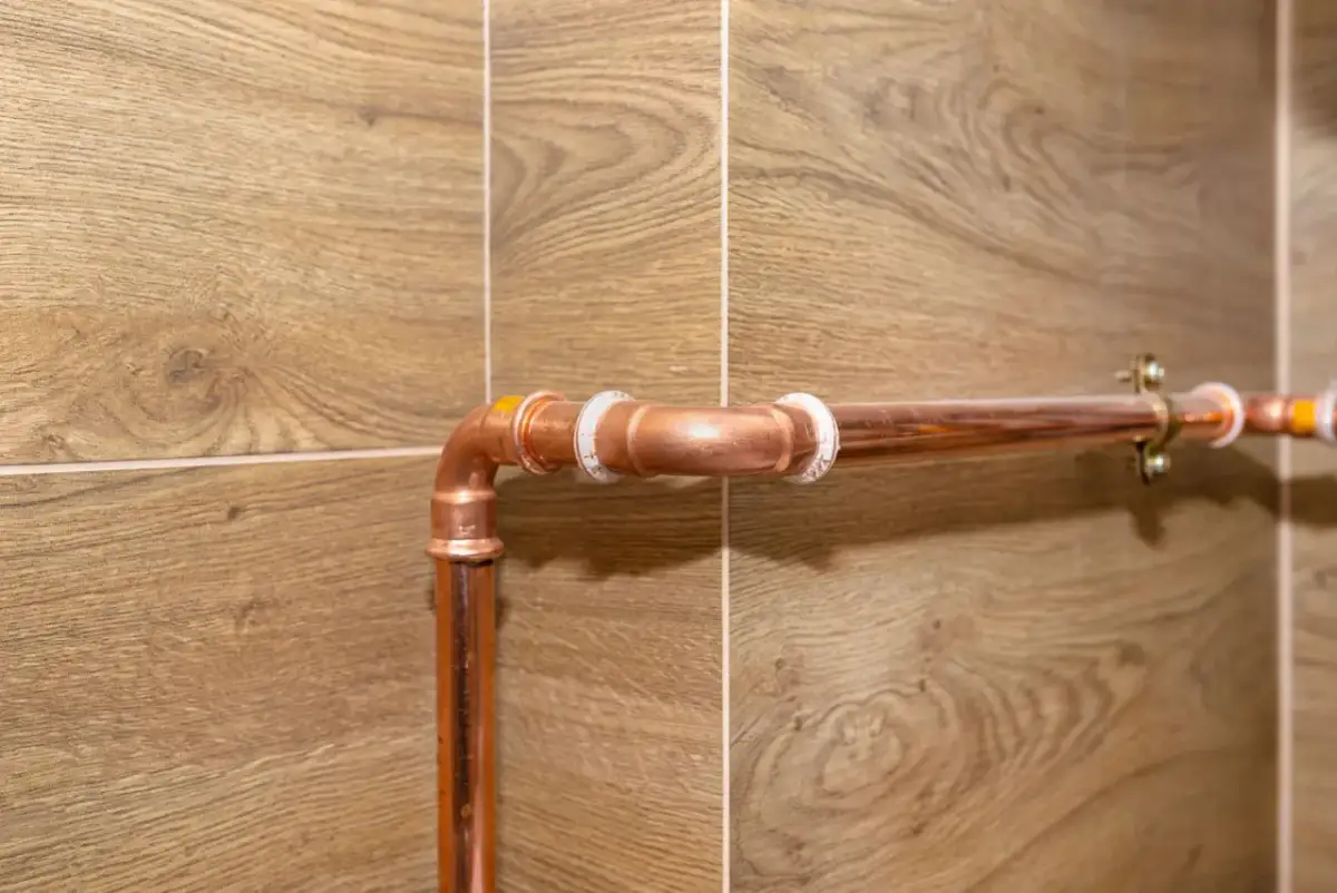 Miedziane rury gazowe zamontowane na ścianie z drewnopodobnymi płytkami. Pokazuje, jak zabudować rury od gazu, aby były estetyczne i bezpieczne.