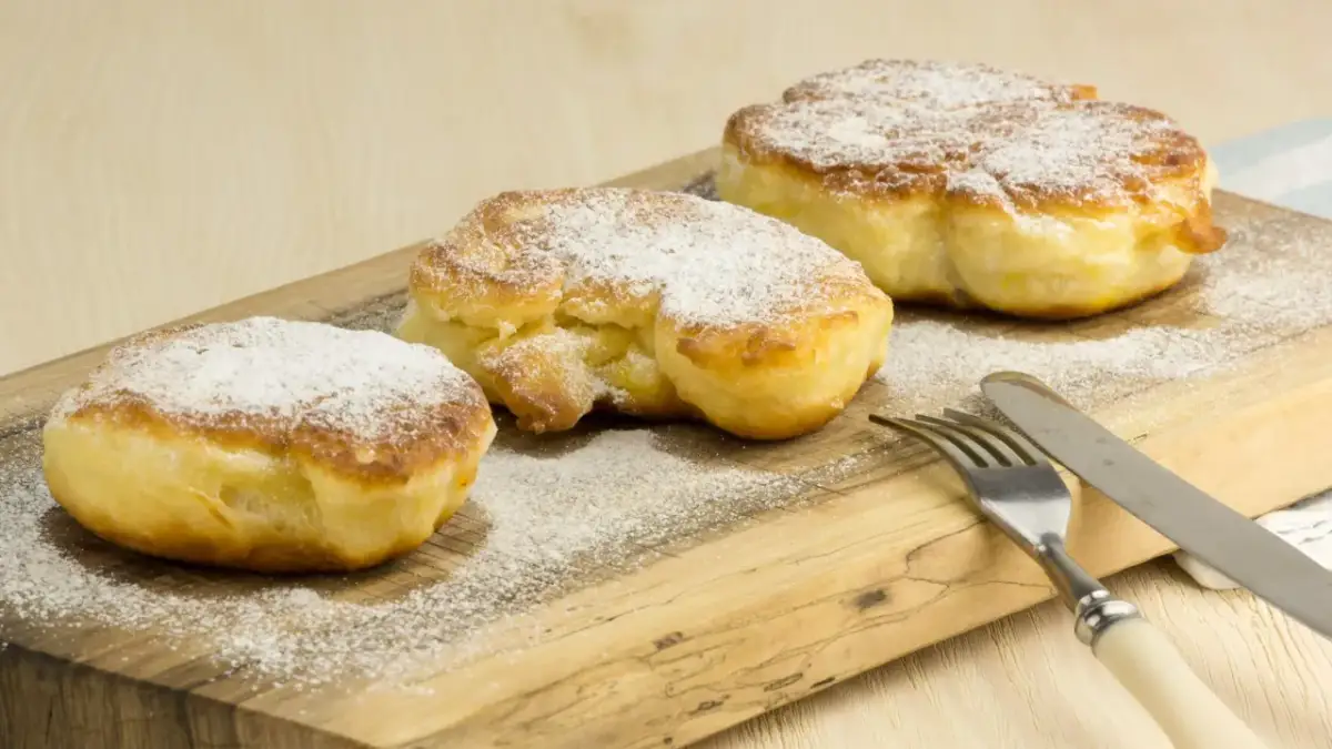 Trzy puszyste racuchy posypane cukrem pudrem na drewnianej desce z widelcem i nożem.