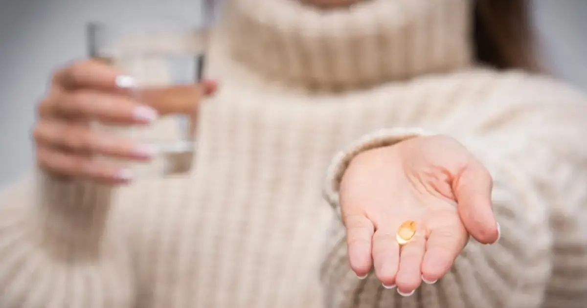 Dłoń trzymająca jedną kapsułkę witaminy B i szklankę wody.