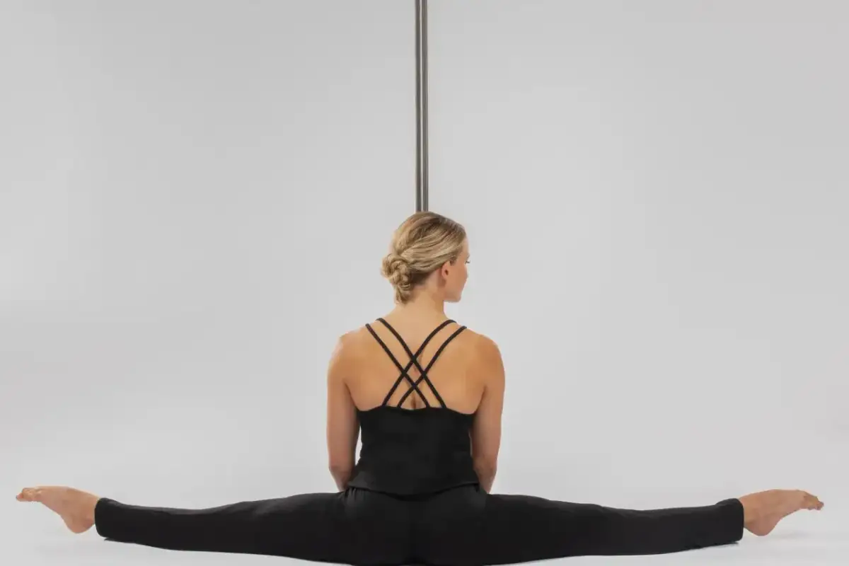 Kobieta w czarnym stroju siedzi w szpagacie przy rurze do pole dance.