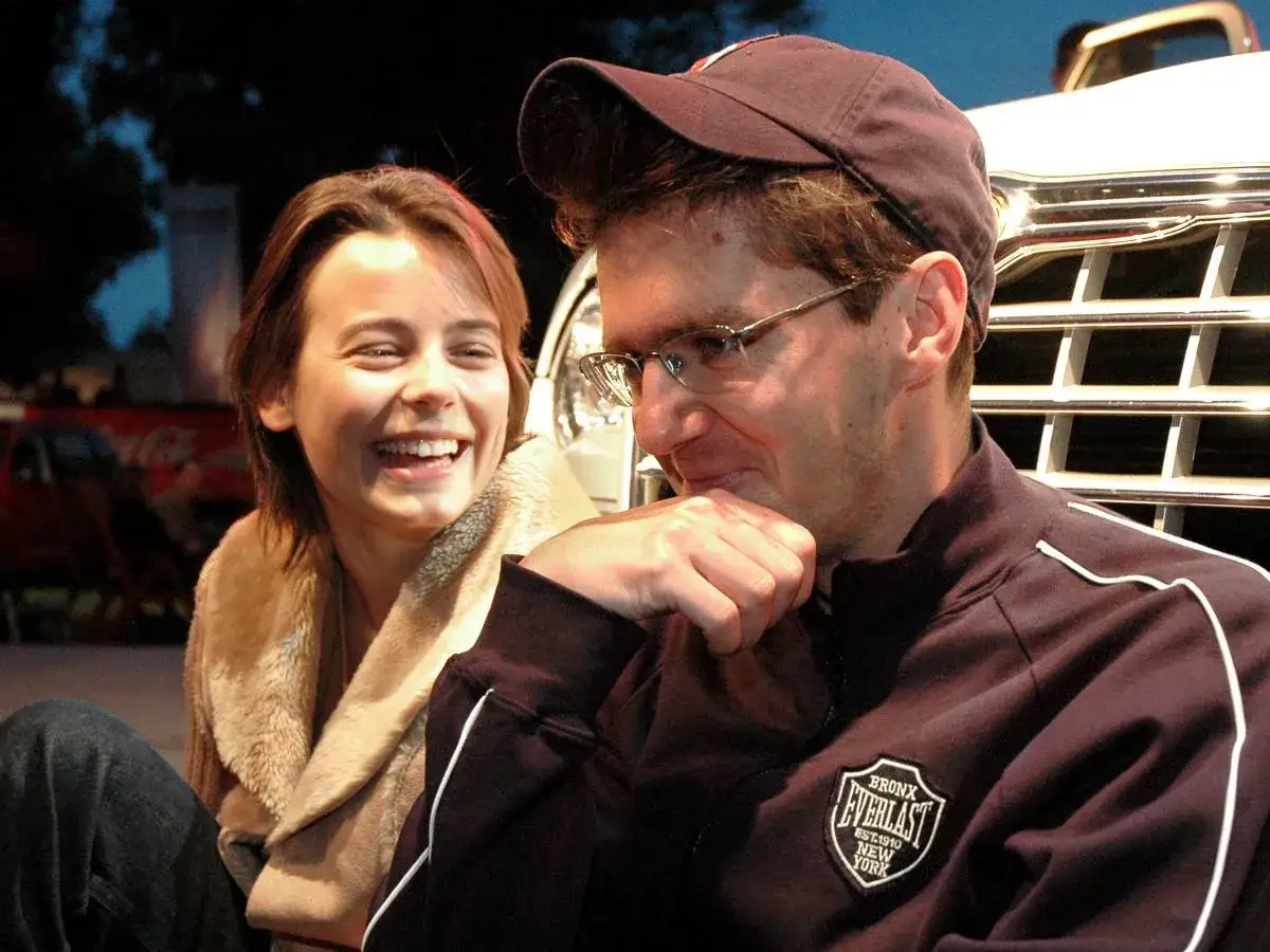 Uśmiechnięta kobieta i mężczyzna w czapce, siedzący obok siebie. Ich rozmowa to o mało co spektakl, pełen śmiechu i emocji.