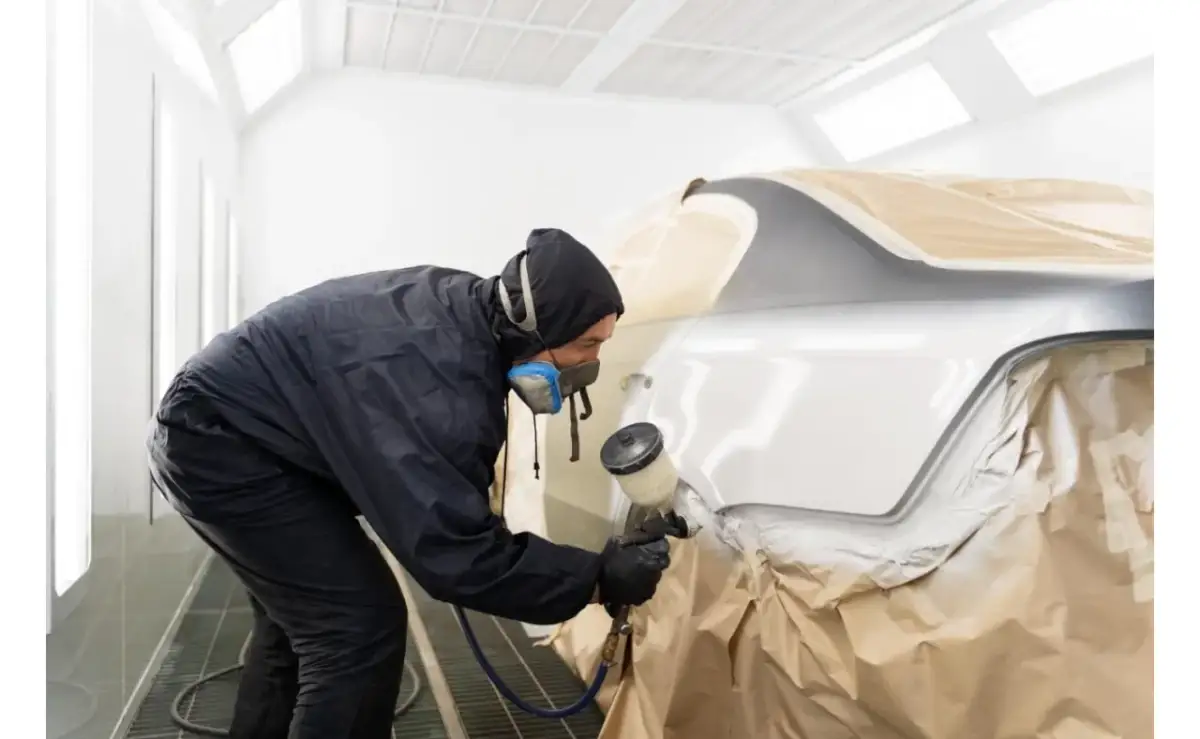 Lakiernik w masce i kombinezonie maluje samochód w kabinie. Czas schnięcia lakieru samochodowego zależy od wielu czynników.