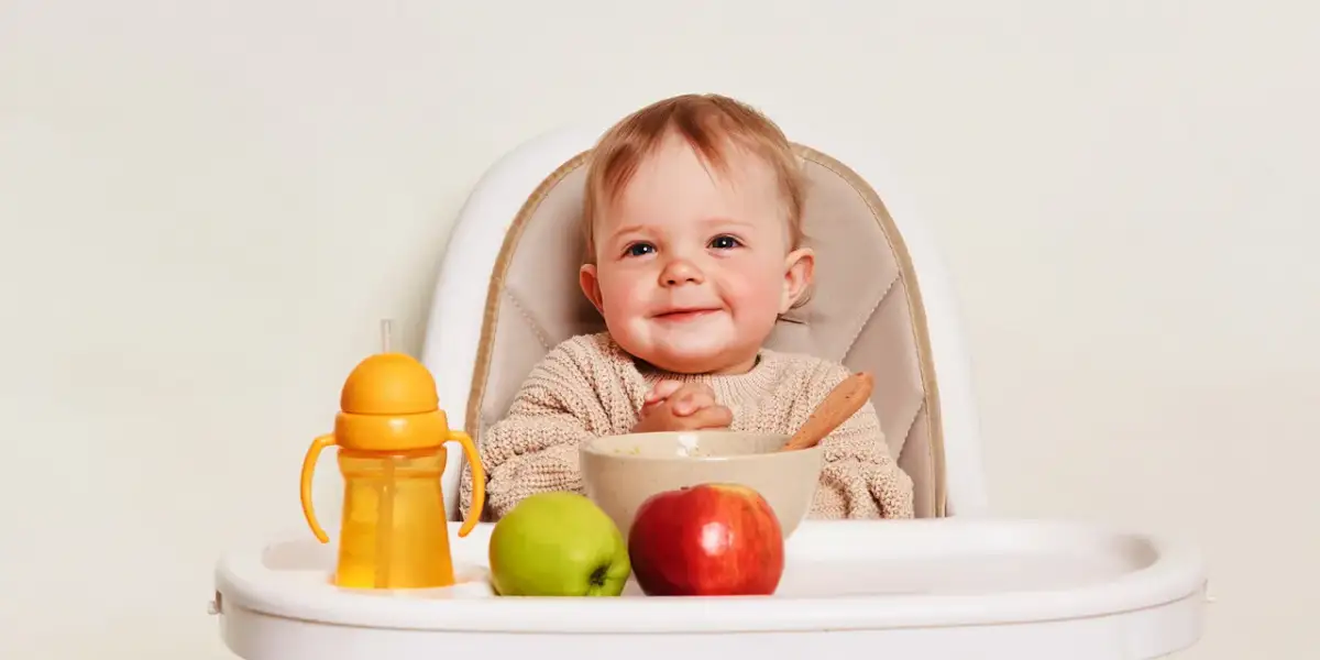 Uśmiechnięte niemowlę w krzesełku, gotowe na pierwsze owoce dla niemowlaka. Obok miseczka, jabłko, gruszka i kubek z uchwytami.