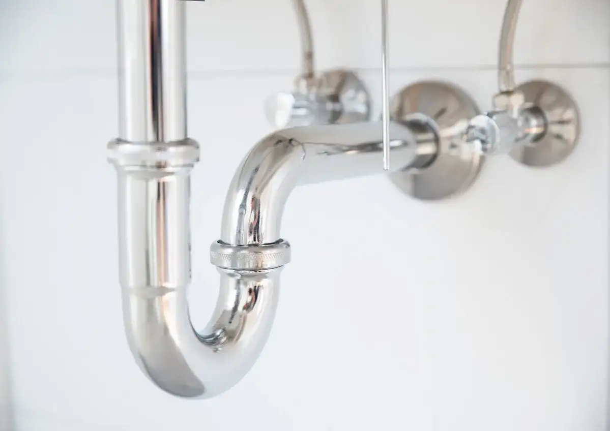 Błyszczący, chromowany syfon do umywalki nablatowej, podłączony do zaworów ściennych.