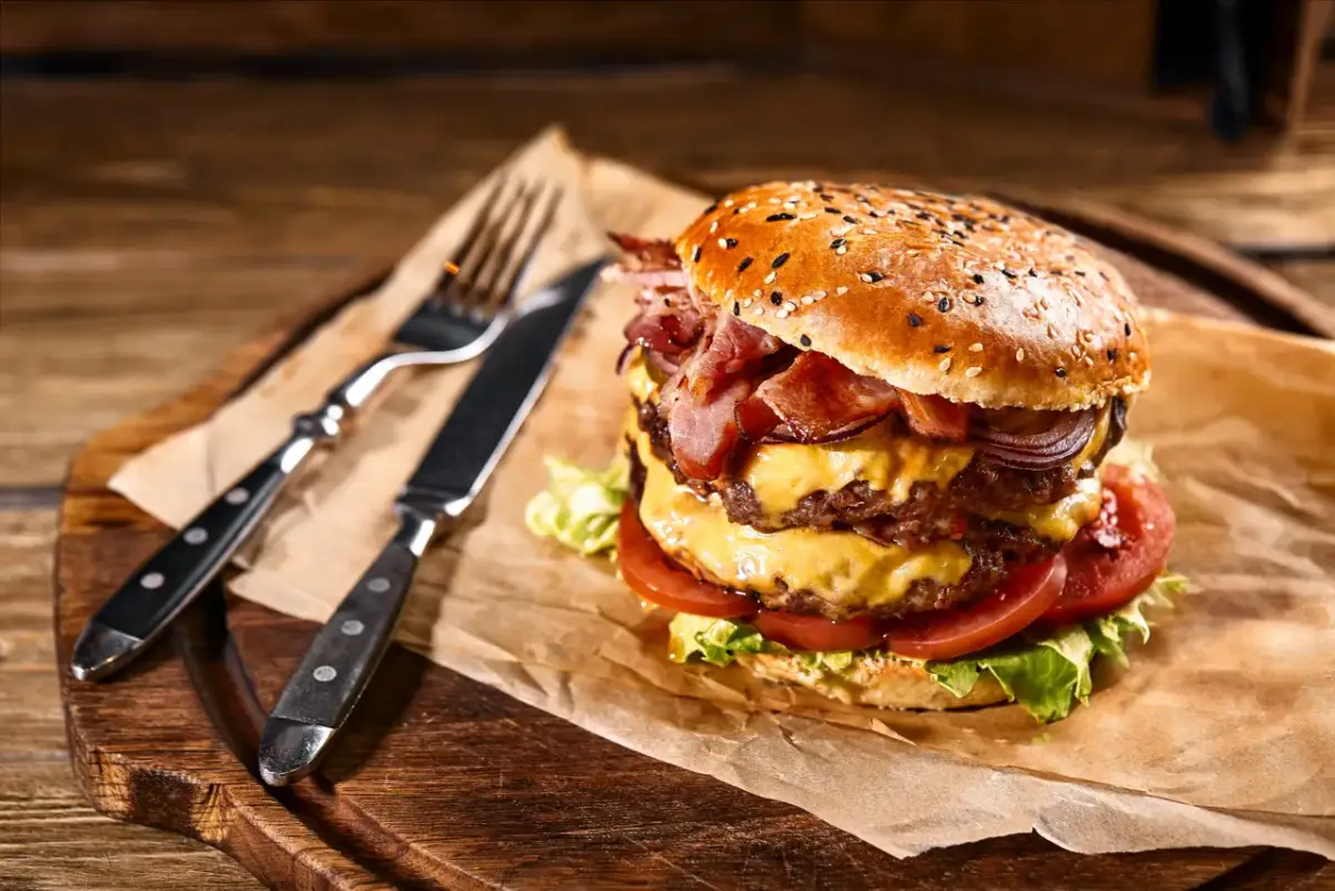 Podwójny burger z bekonem, serem, pomidorami i sałatą na papierze, obok sztućców na desce.