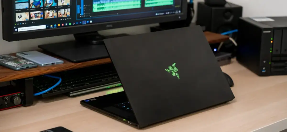 Czarny laptop Razer z podświetlanym logo, obok monitor z edytorem wideo i dysk sieciowy. Prawdopodobnie najdroższy komputer gamingowy na świecie.