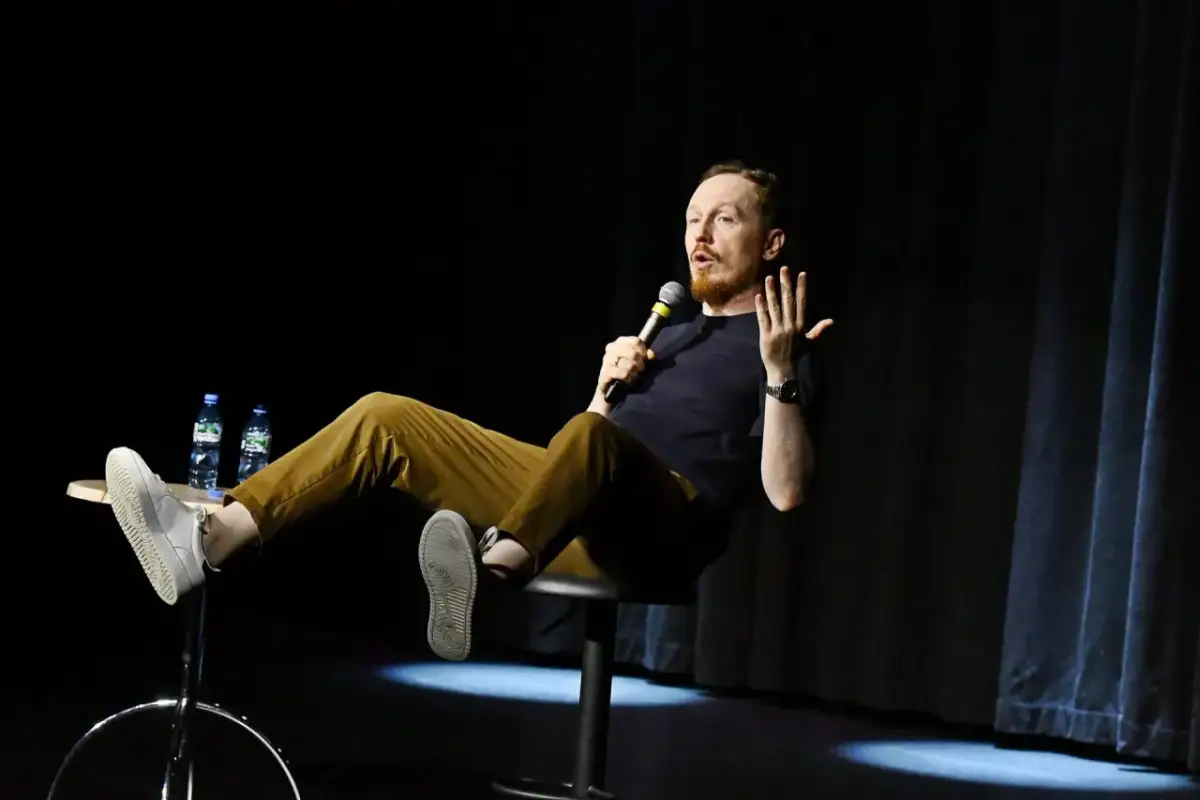 Bartek Toczek w swoim stand-upie opowiada dowcipy, siedząc na stołku i trzymając mikrofon.