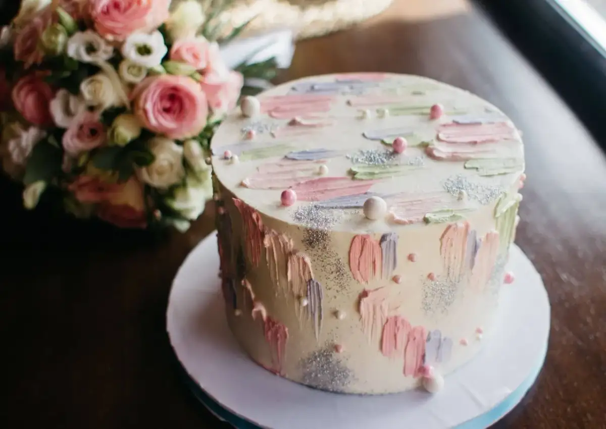 Okrągły tort z pastelowymi dekoracjami i bukietem róż w tle.