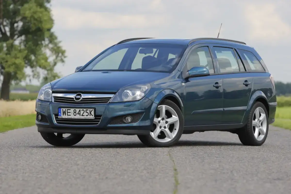 Granatowy Opel Astra Cosmo w wersji kombi stoi na asfaltowej drodze, otoczony zielenią i drzewami.