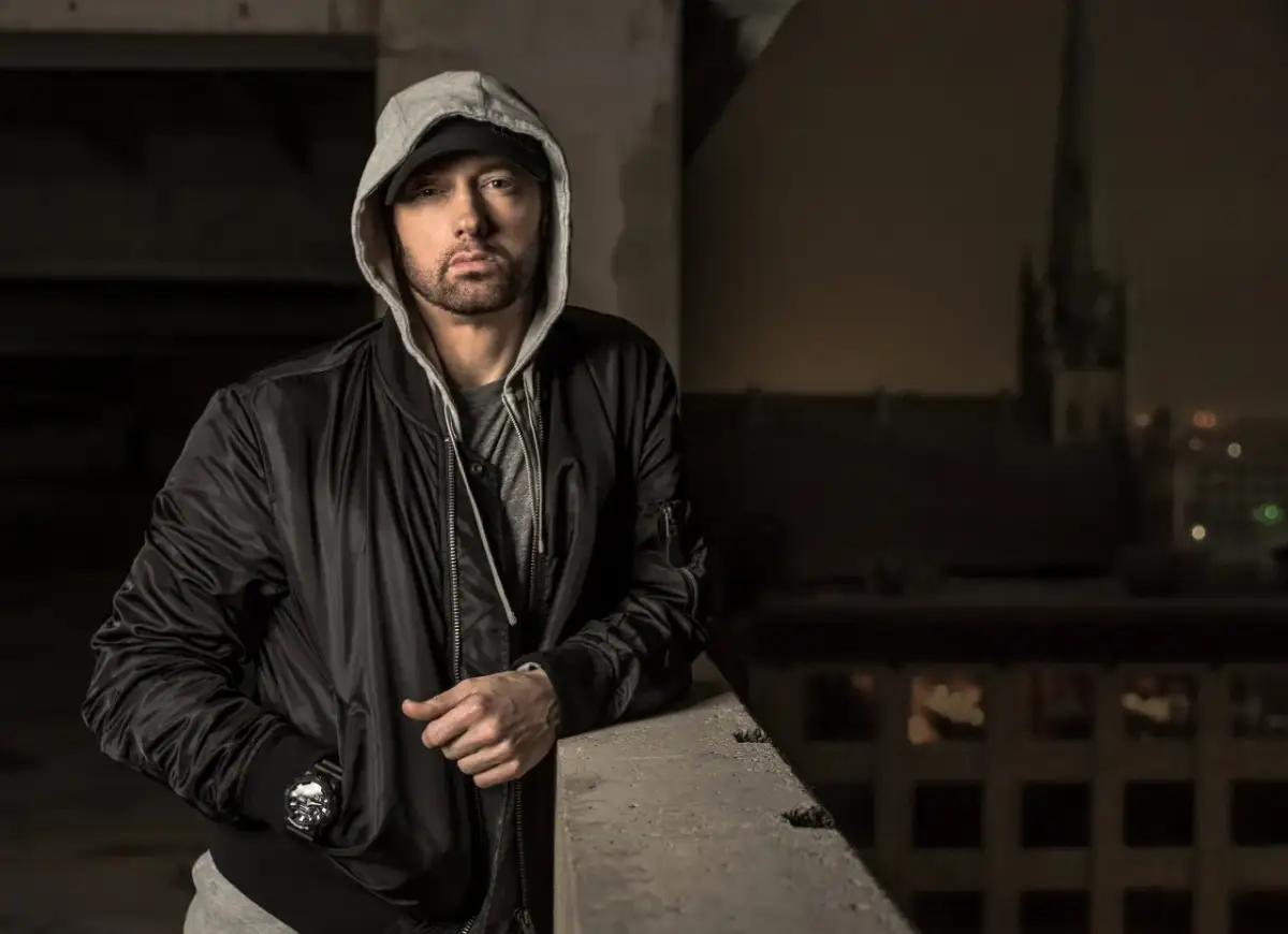 Eminem w czarnej kurtce bomberce i szarej bluzie z kapturem, oparty o betonową barierkę.